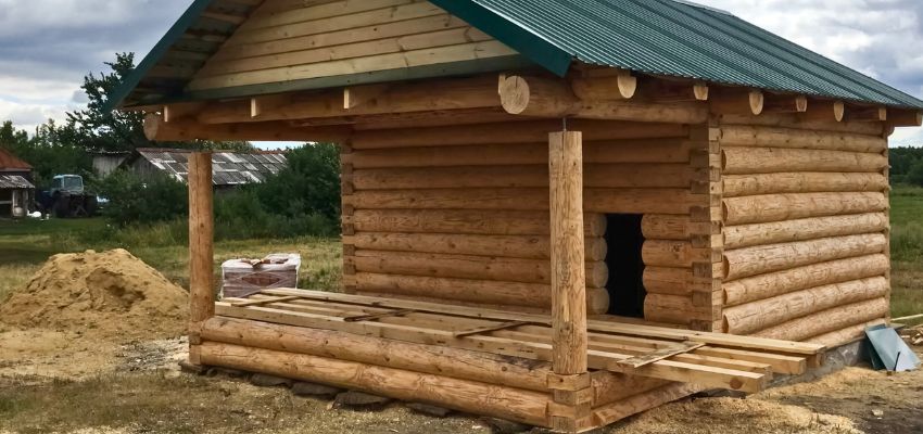 Partially assembled small log cabin made from an affordable log cabin kit under 5000, showing exposed logs, front porch framing, and green metal roof during construction.