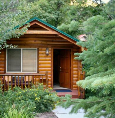 Charming finished log cabin with green roof, wooden front door open, and cozy porch surrounded by pine trees and flowers – an inviting example of log cabin kits under 5000 fully assembled.