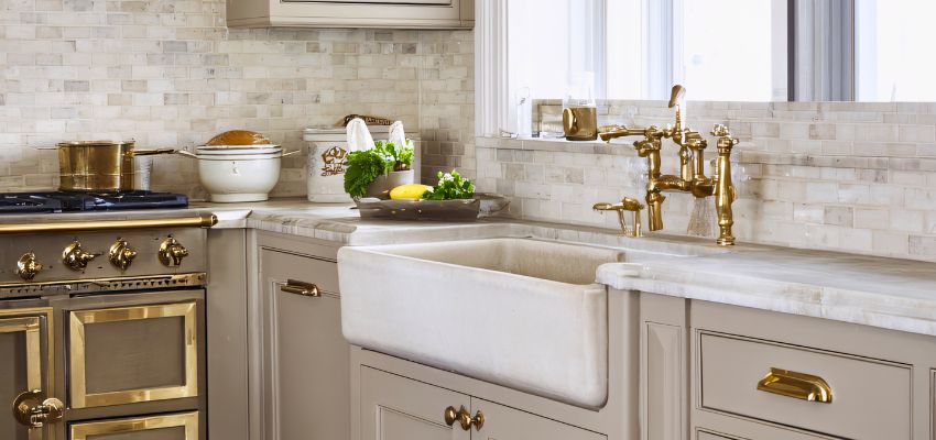 Elegant white farmhouse sink integrated into a marble countertop with luxurious brass faucet, surrounded by neutral cabinetry and a professional brass-accented stove, showcasing premium farmhouse sinks in modern kitchens.