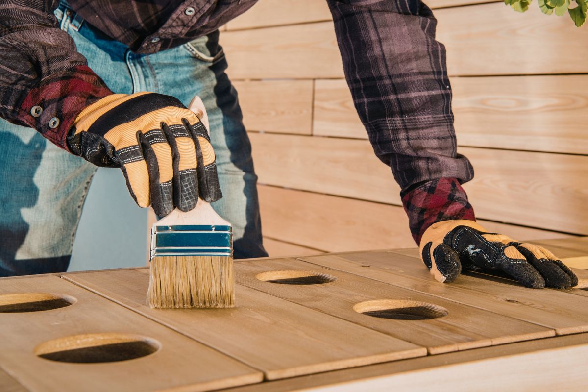 Person wearing gloves applying wood sealer with a brush on an outdoor wooden project