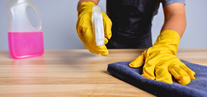 Person wearing yellow gloves spraying and wiping a wooden table with a cloth, demonstrating how to clean a sticky wooden table safely.