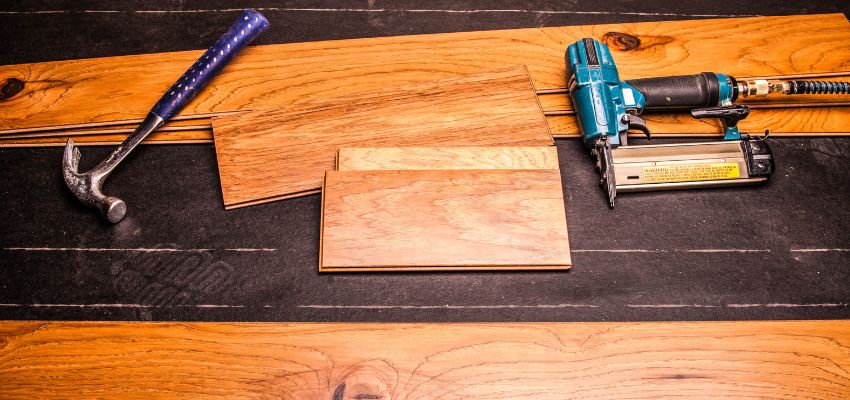 Close-up of unfinished hickory wood flooring planks laid out on a workbench with a hammer, nail gun, and staples, ready for professional hardwood installation.